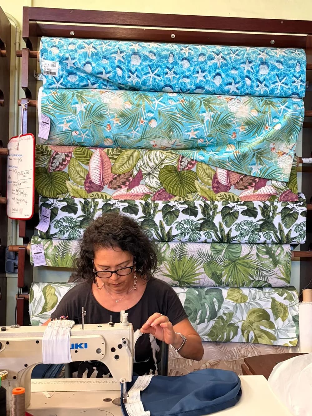 Couturière à l'œuvre dans l'atelier de confection Couleur Fenua, devant une sélection de tissus d'ameublement